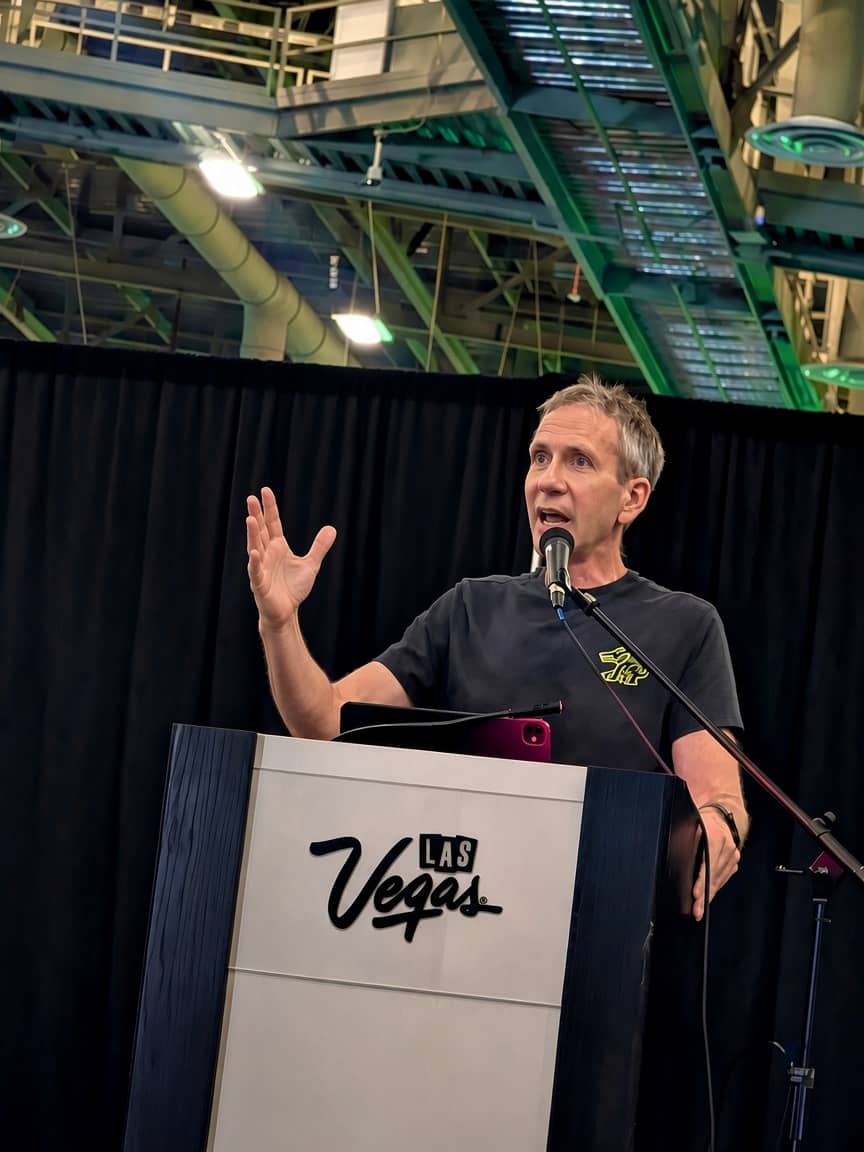 Jeff Crume mid-sentence at a lectern branded with the Las Vegas script logo, one hand raised to make a point, the convention hall's exposed green steelwork above him